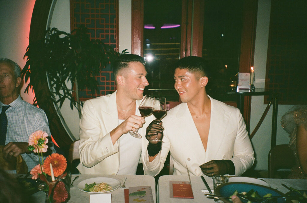 Film photo of grooms clinking red wine glasses and smiling at each other.