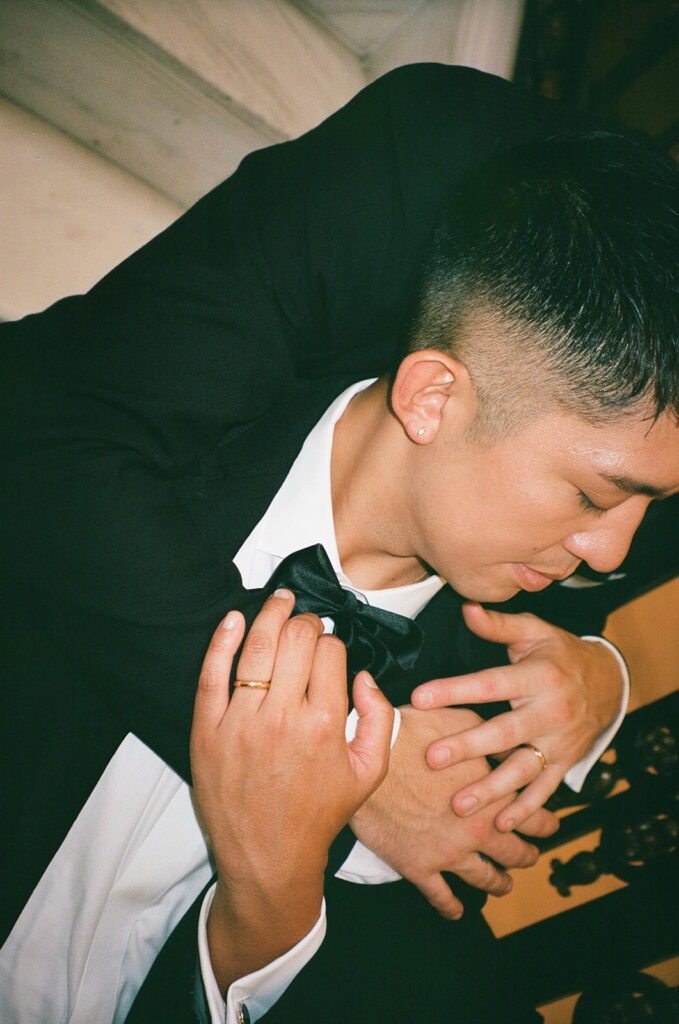 Groom holding other groom from behind and them holding arms, gold wedding bands on display.