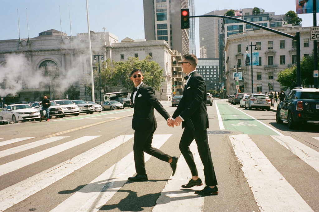 Men in formal black wedding suits holding hands walking across a crosswalk.