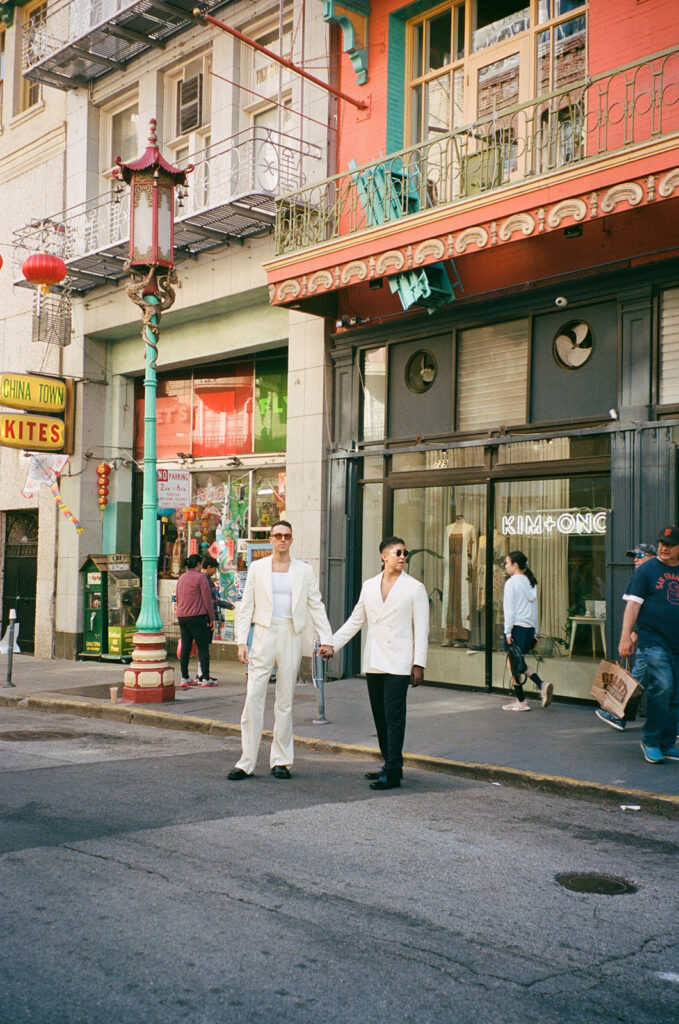 From across the street, two grooms holding hands looking in different directions.