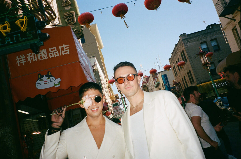 Photo from below with red Chinese lanterns above two men, one with a pink rose over one eye and one with orange lens sunglasses.