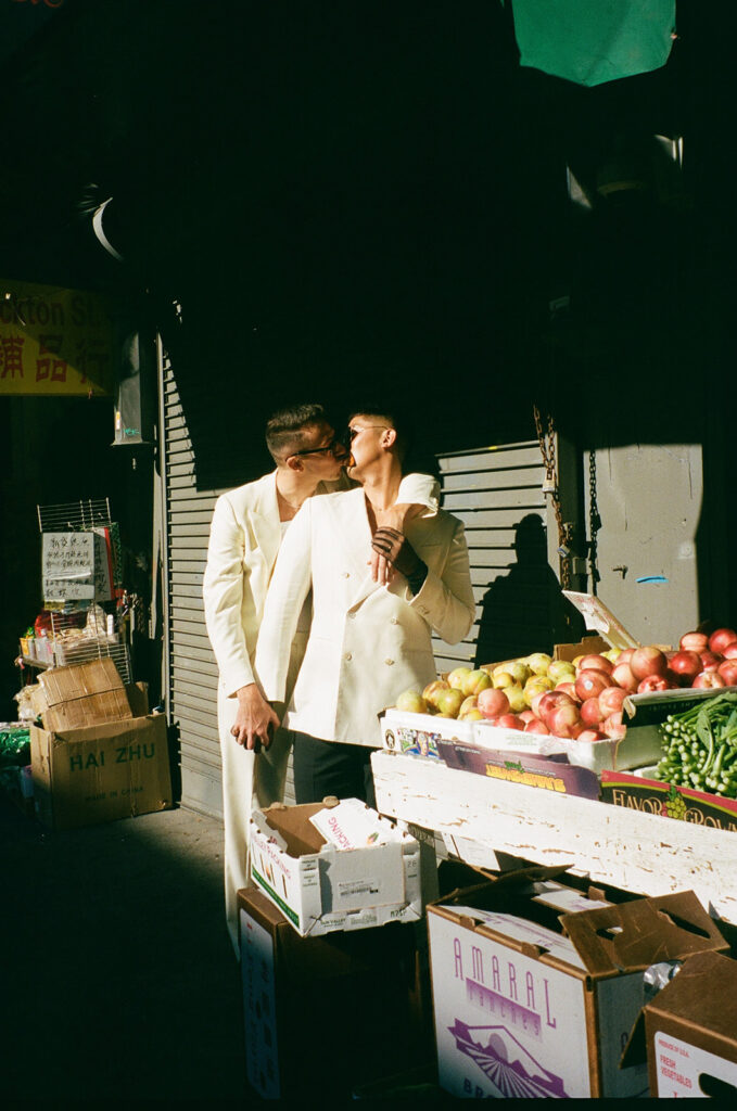 Men kissing in front of a fresh produce in the street.