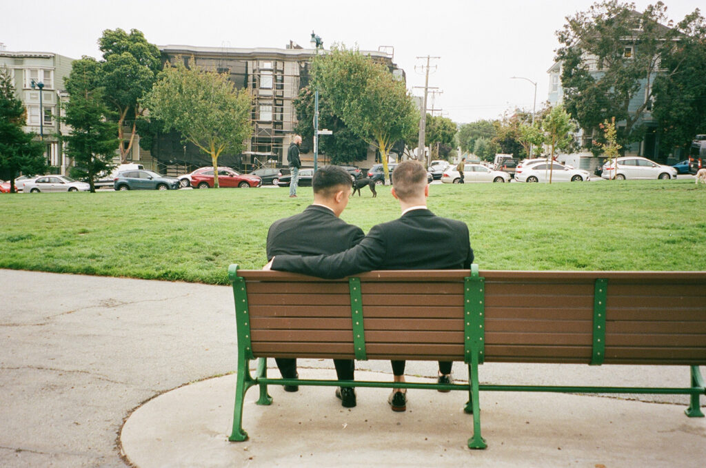 Film photo of gooms in formal suits sitting on a bench in the park from behind with one wrapped arm around the other.