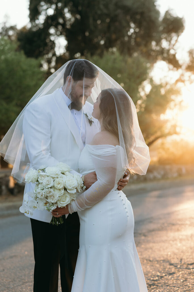 Veil over both the bride and groom and she holds her white rose bouquet as the sunsets behind them.
