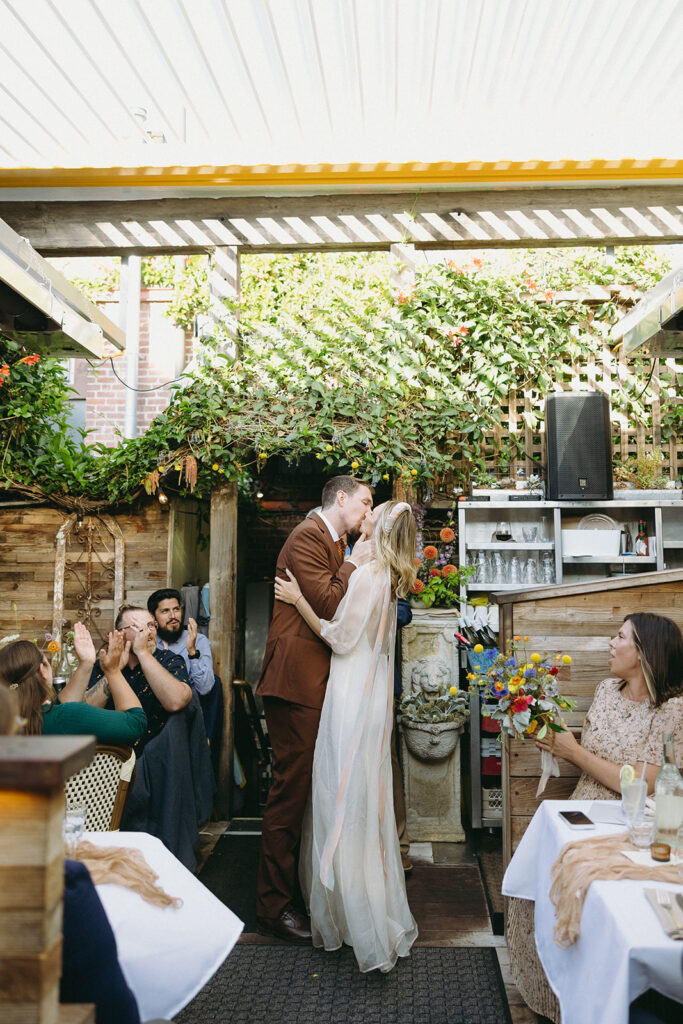 Wedding ceremony kiss of groom in a rust colored suit and bride in a flowy gown and headband strings flowing down her back.