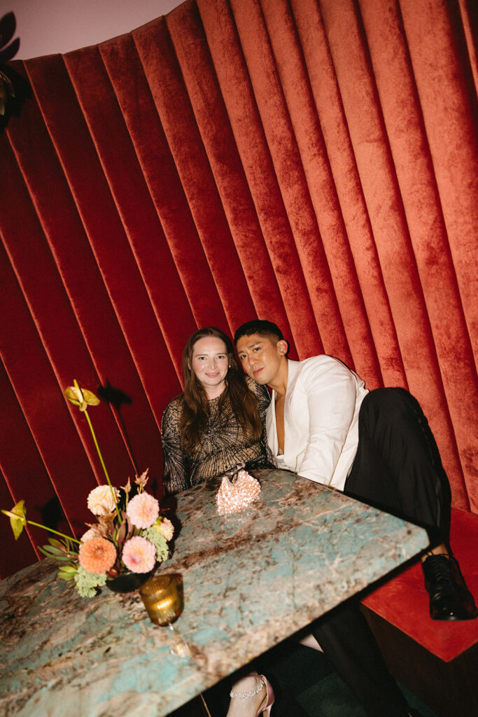 Groom and friend sitting in red velvet booth with a green marble table and floral centerpieces.