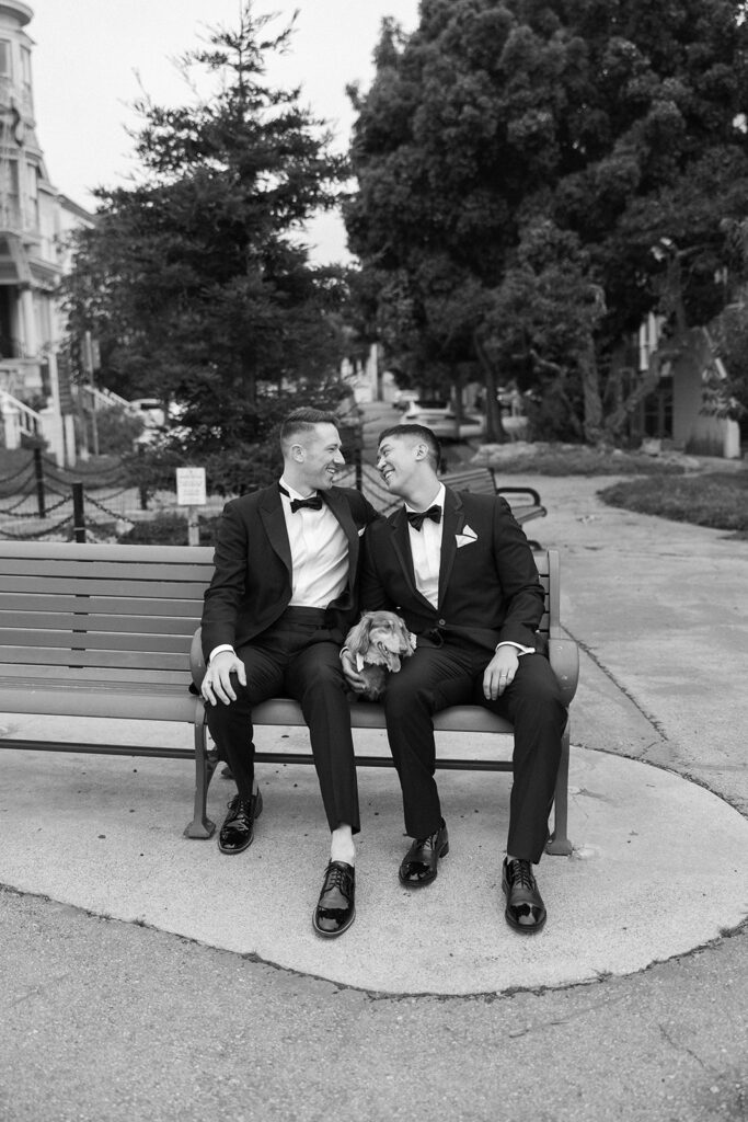 Grooms in formal suits sitting on a bench in the park with their dachshund.
