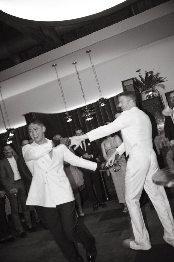 Grooms, one in all white and the other in white suit jacket and black trousers, holding hands dancing.