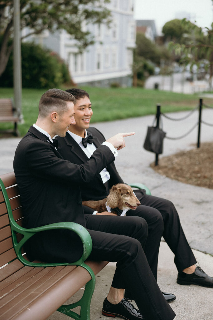 Grooms in formal suits sitting on a bench in the park with their dachshund, one pointing to look somewhere.