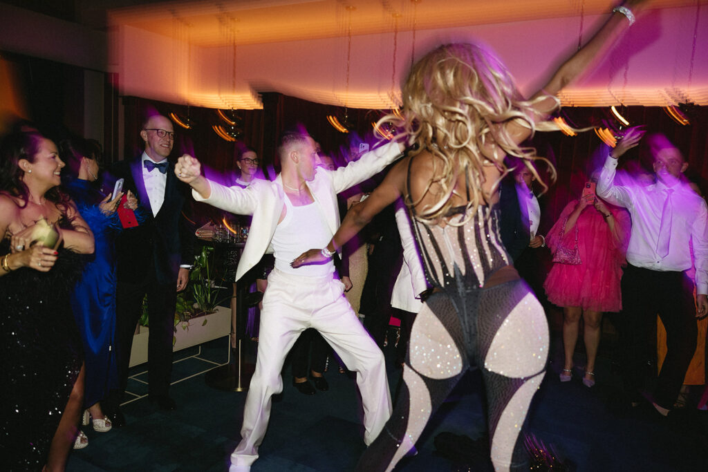 Drag performer in bedazzled one piece on the middle of the wedding dancefloor with groom wearing white.