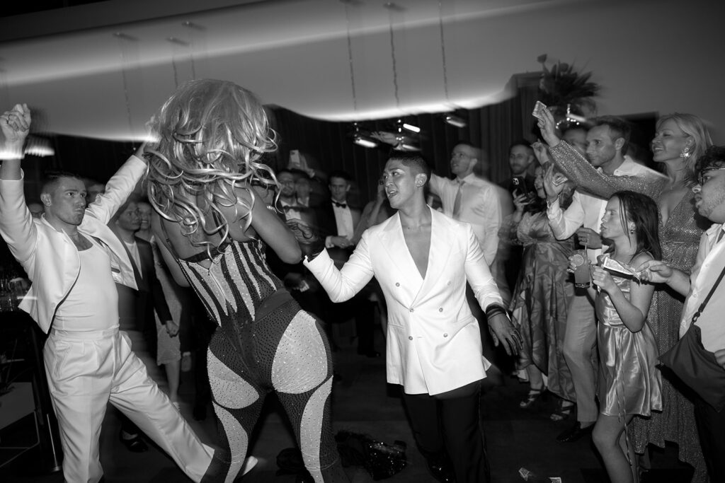 Drag performer in bedazzled one piece on the middle of the wedding dancefloor with both grooms wearing white.