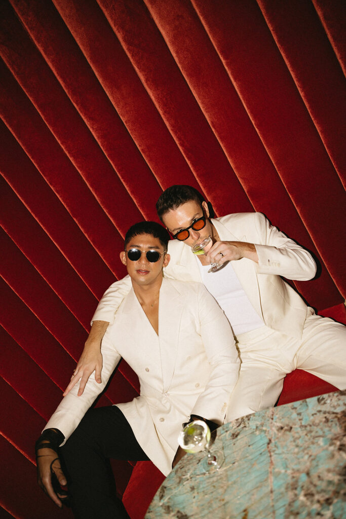 Two grooms sitting in a red velvet booth wearing black and white and sunglasses with martinis.