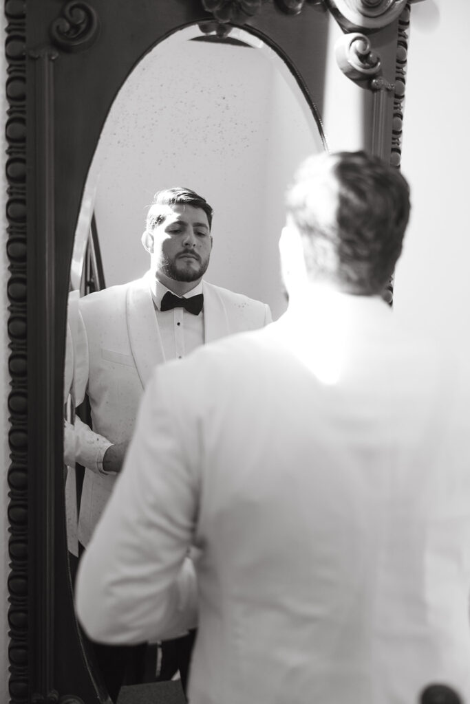 Groom looking at himself in the mirror as he button his white suit jacket.