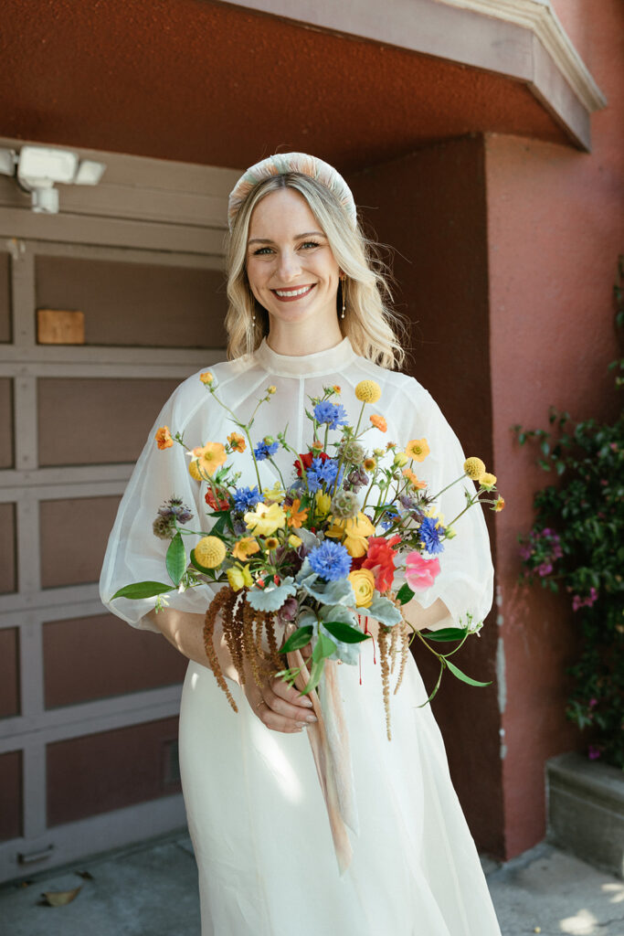 Bridal portrait with a chunky headband in short curled blonde hair and long dangly earrings and a high neck wedding gown with a large wildflower bouquet.