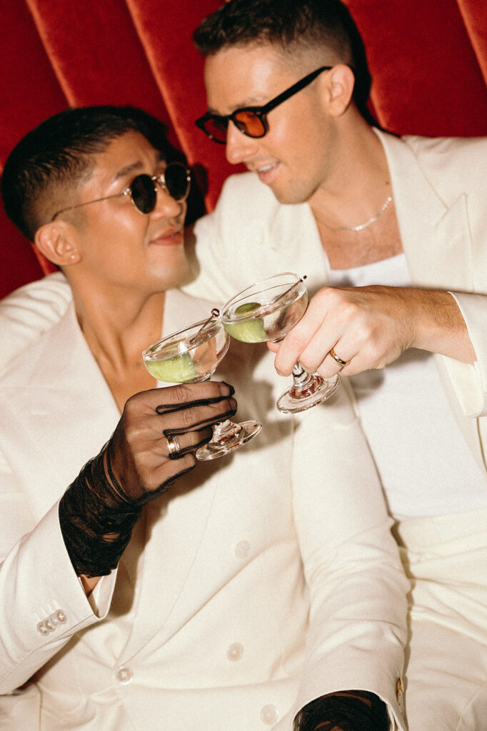Grooms cheersing their martinis with an olive while one has a mesh black glove on and both are wearing sunglasses.