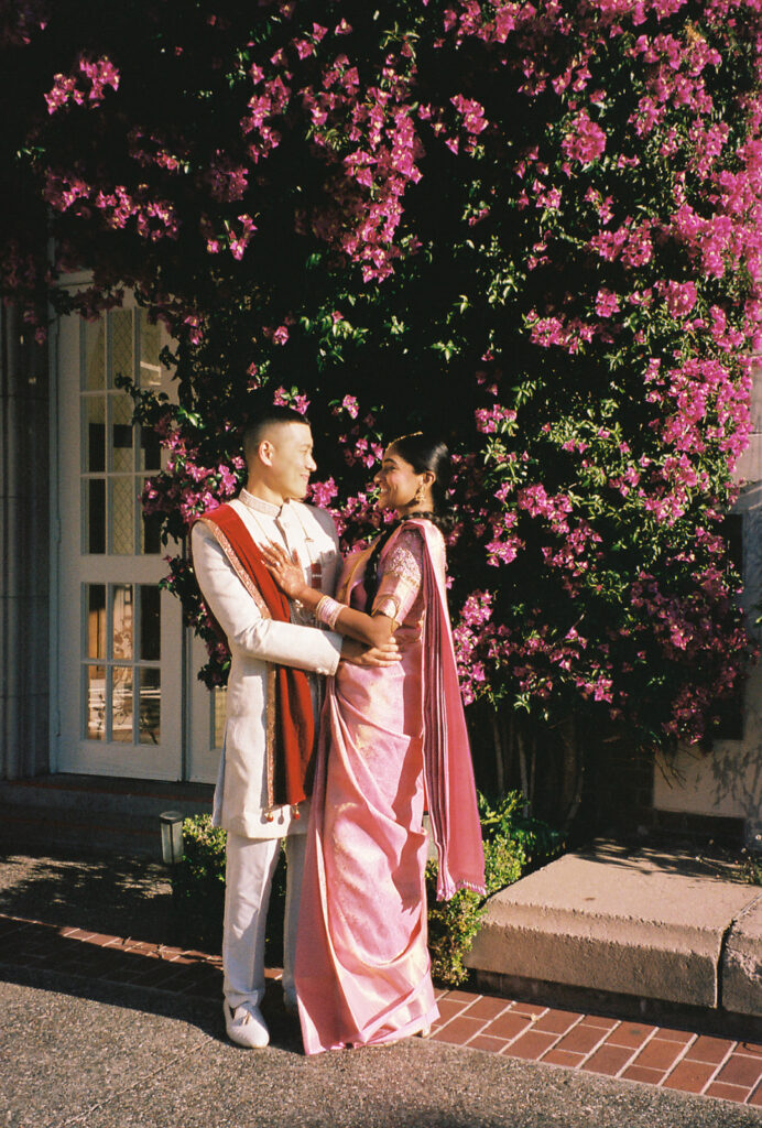 Film photo of bride, in a pink saree, and groom, in a white sherwani and red dupatta, holding each other and smiling at each other.