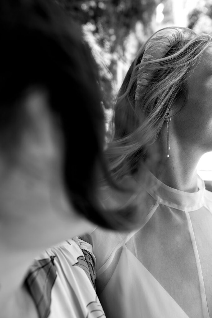 Blurry foreground and bridal hair and jewelry in the background in focus. Chunky headband in short curled blonde hair and long dangly earrings and a high neck wedding gown.