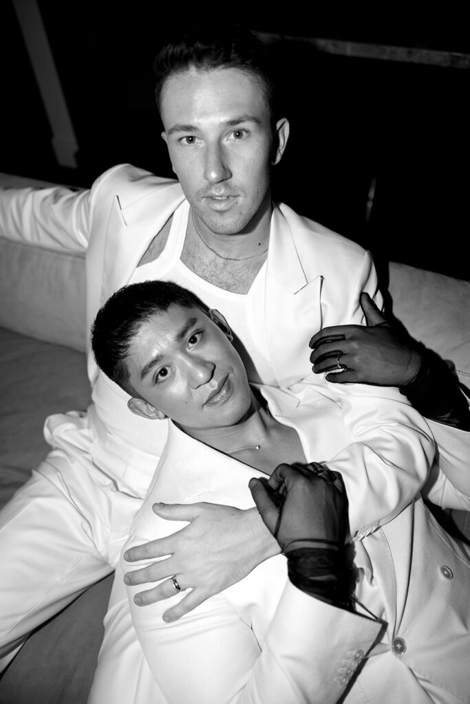 Grooms dressed in all white, one laying across the others chest, both looking lovingly up at the camera.