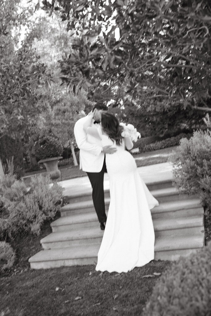 Bride kissing groom from a lower step on a set of stairs outside.