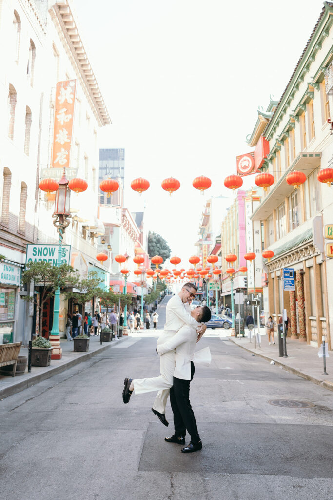 Man lifting up another man dressed in all white in the streets of Chinatown with red lanterns above them.