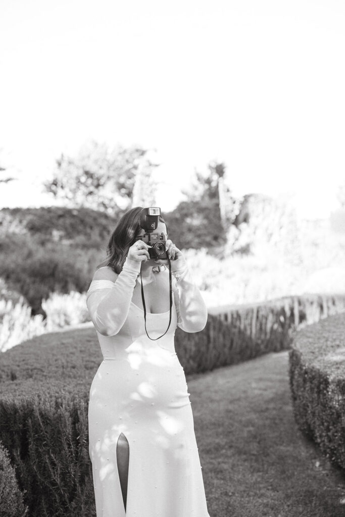 Bride in a fitted wedding gown with sheer long sleeves holding a film camera up to her face to take a photo.