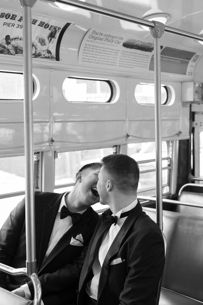 Two grooms laughing closely while sitting on the SF trolley in their suits.