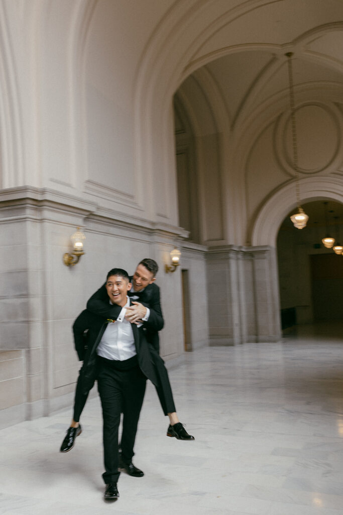 One groom carrying the other through the halls of SF City Hall laughing.