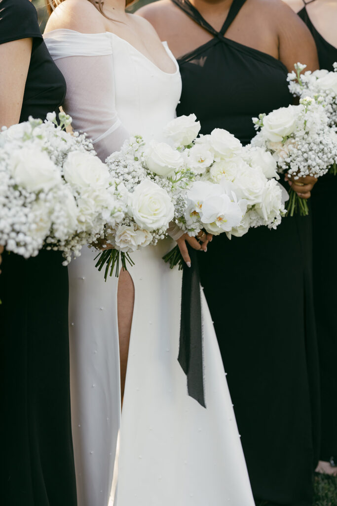 Bridesmaids, in all black bridesmaid dresses, holding their all white rose and babys breath bouquets next to the bridal bouquet.