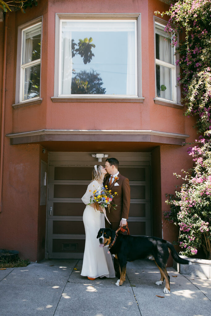 Bride and groom standing in front of SF red building and brown garage door with their large dog. 
