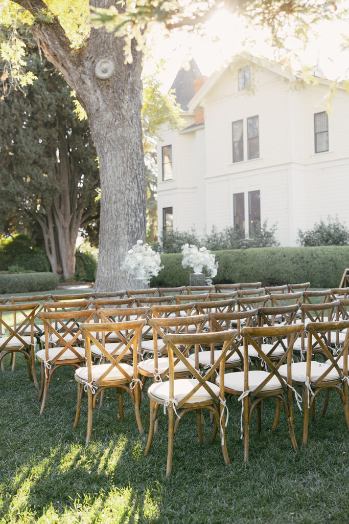 Wooden chairs with beige cushions for outdoor ceremony under a tree.