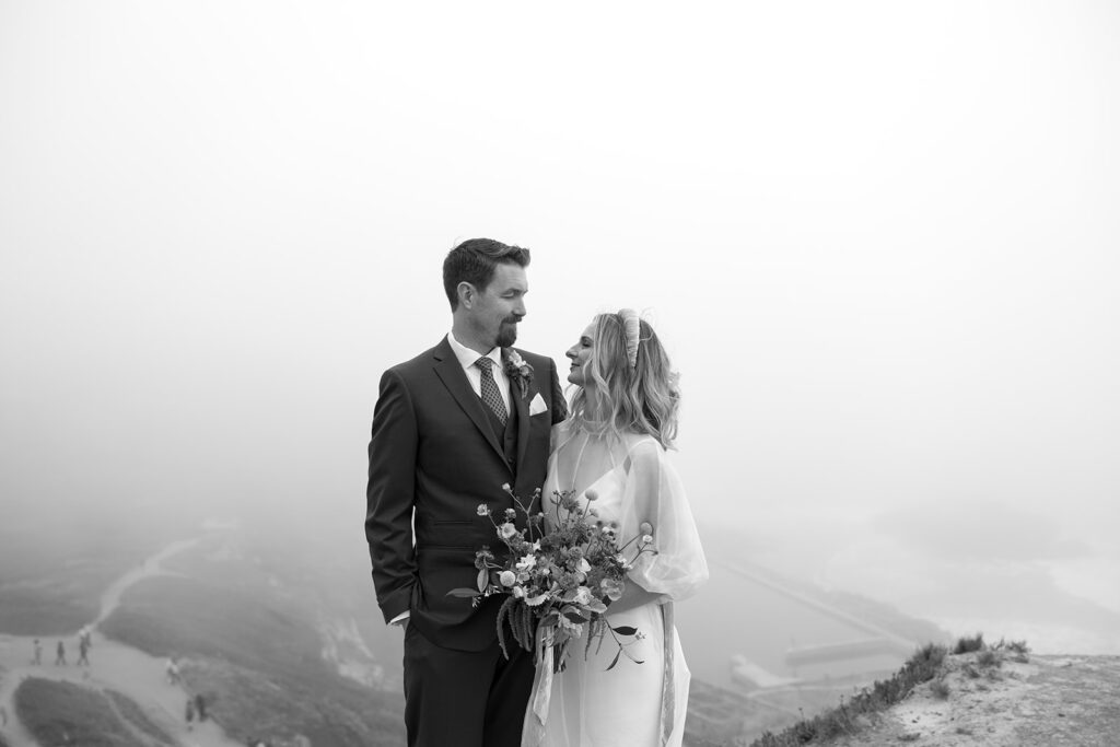 Black and white photo of a bride and groom standing on a cliffside staring lovingly at one another. Wildflower bouquet and bride has short hair with a chunky headband.