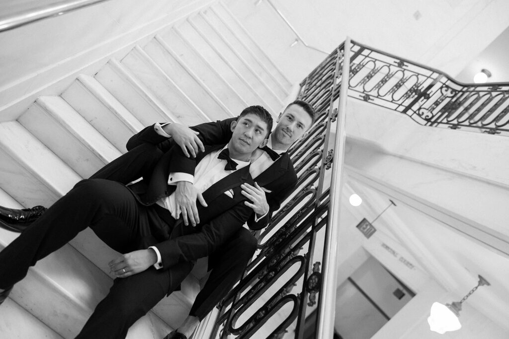 Two grooms sitting on staircase in formal wedding suits holding each other.
