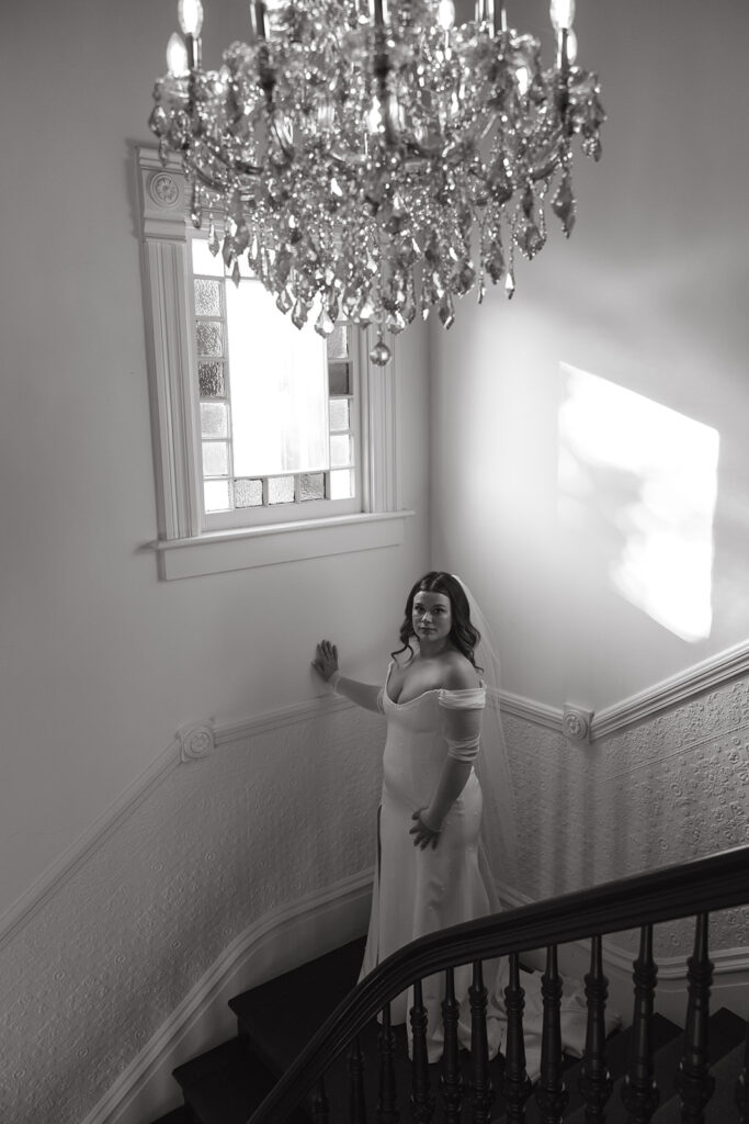 Bride in stairwell under a chandelier and holding hand on the wall under a stained glass window.