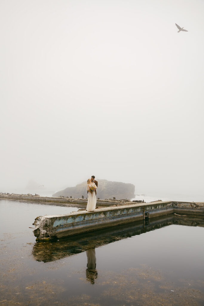Bride and groom in the distance standing on a gloomy broken dock embracing one another.