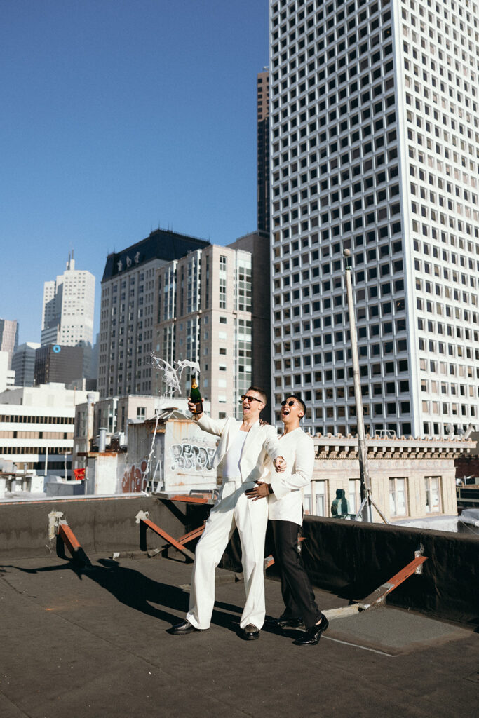 Grooms on rooftop popping open a bottle of champagne.