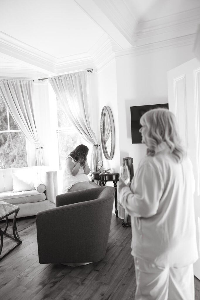 Bride sitting at a vanity getting ready while mother of the bride drinks champagne in foreground of the photo.