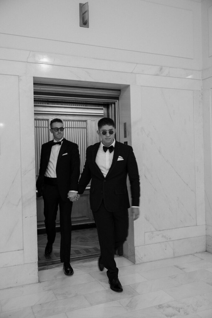 One man leading another out of the elevator holding hands, wearing formal suits.