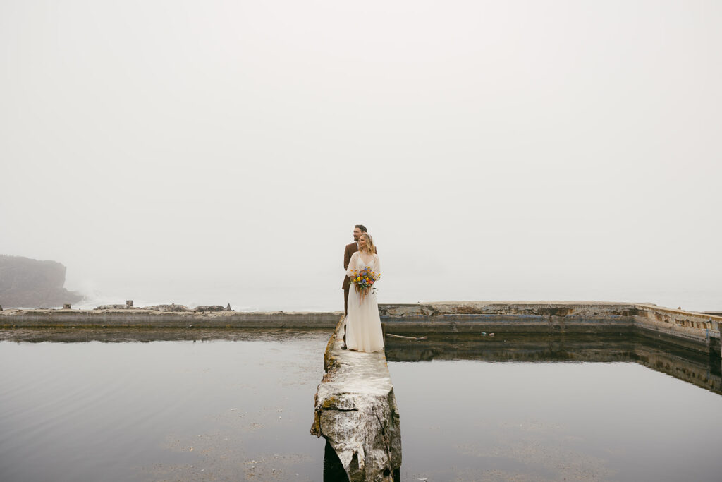 Bride and groom in the distance standing on a gloomy broken dock embracing one another.