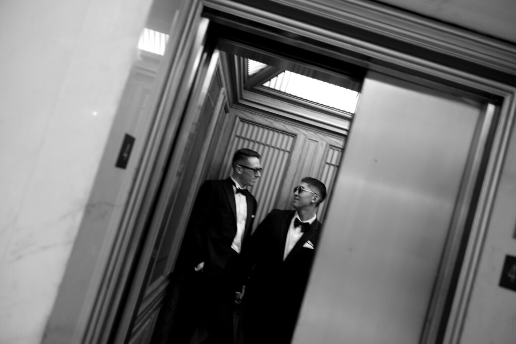 Editorial elevator photo of two grooms in formal suits closed mouth smiling at each other.
