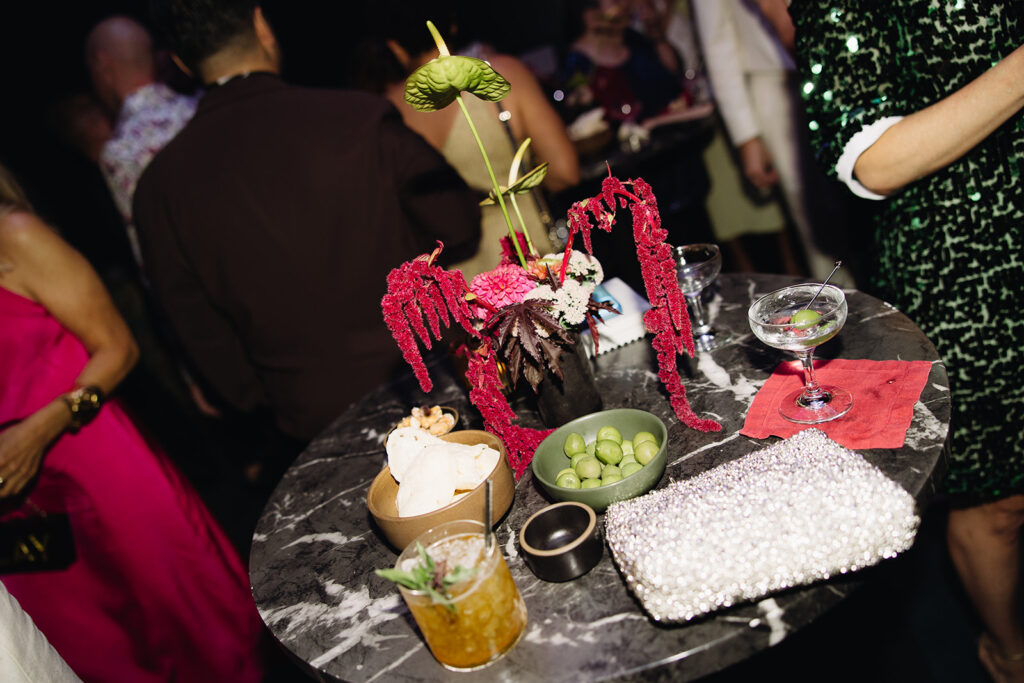 Wedding cocktail table with modern floral arrangement, silver sequin purse, cocktail, and olives.