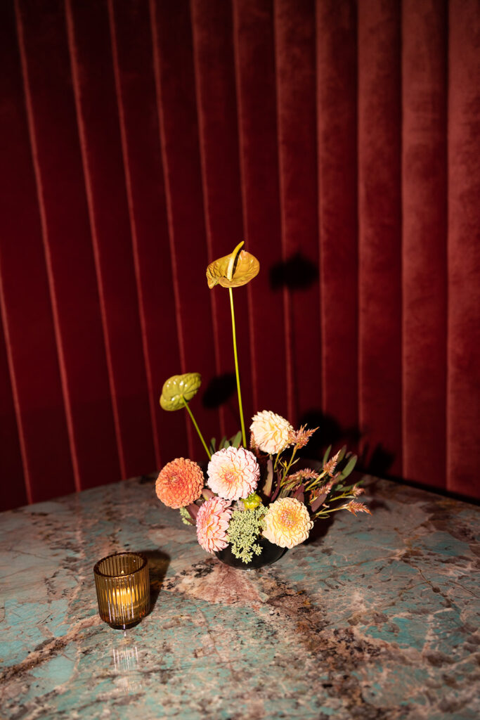 Layered small floral arrangement with dahlias and anthurium on a green and brown marble table at a red velvet booth.