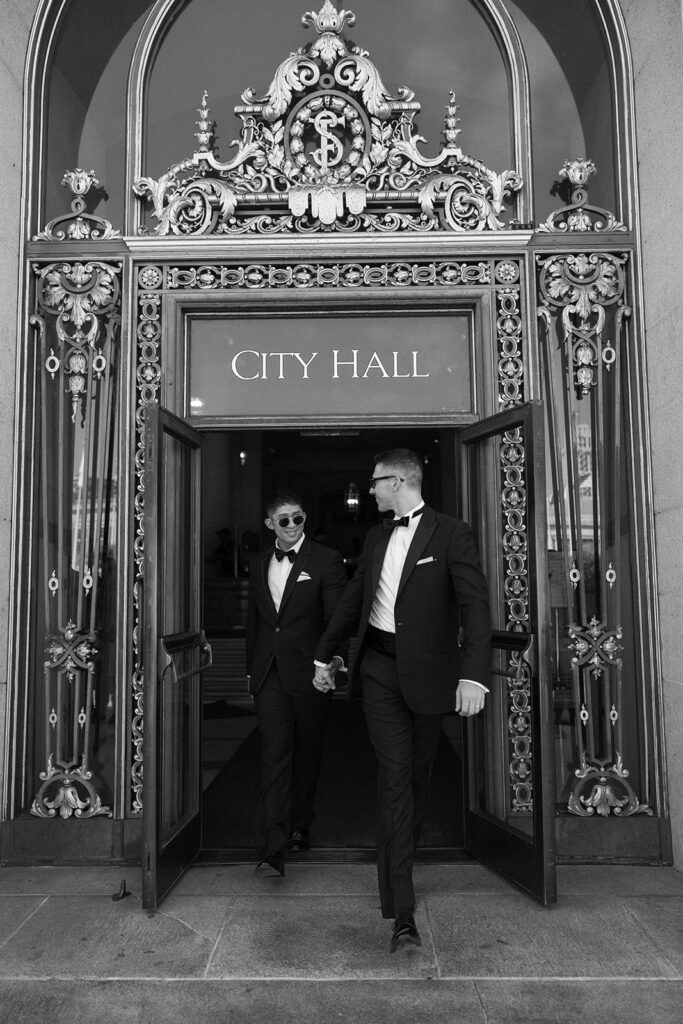 One groom leading the other out of city hall smiling back at him.