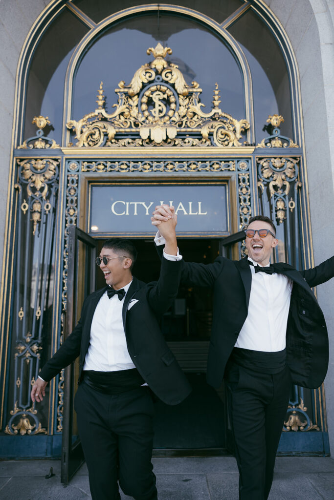 Two gay men running out of city hall cheering with their arms up celebrating their marriage.