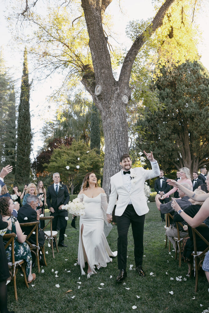 Couple walking back down the aisle laughing and cheering while guests throw white rose petals.