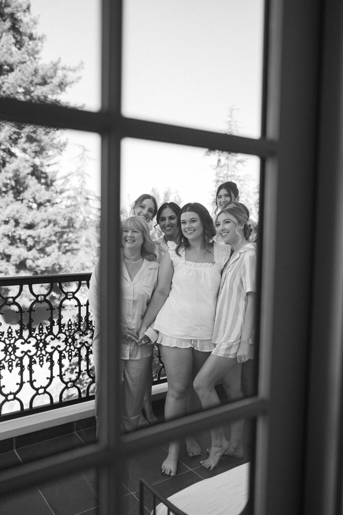 Captured through a window, the bridal party in their morning pajamas and finished hair and makeup smiling outside on the balcony.