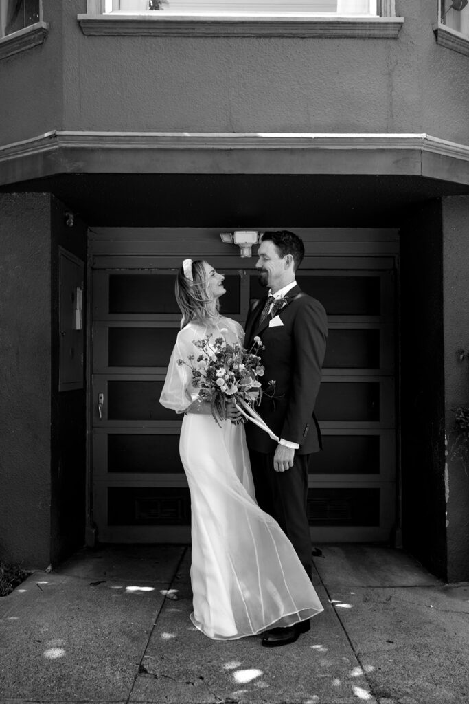 Black and white photo of bride and groom standing in front of garage door with wind blowing in brides short hair. Bride is holding a large wildflower bouquet.