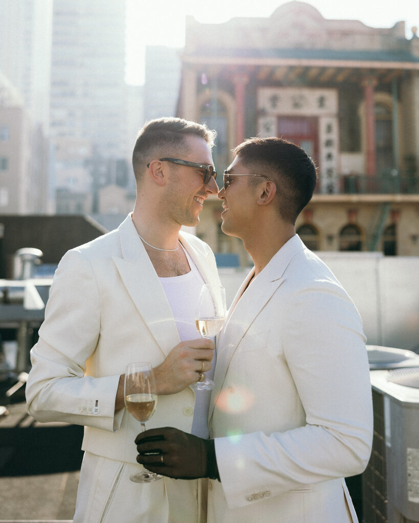 With Chinatown in the background, two grooms in white suits holding wine glasses touching noses.