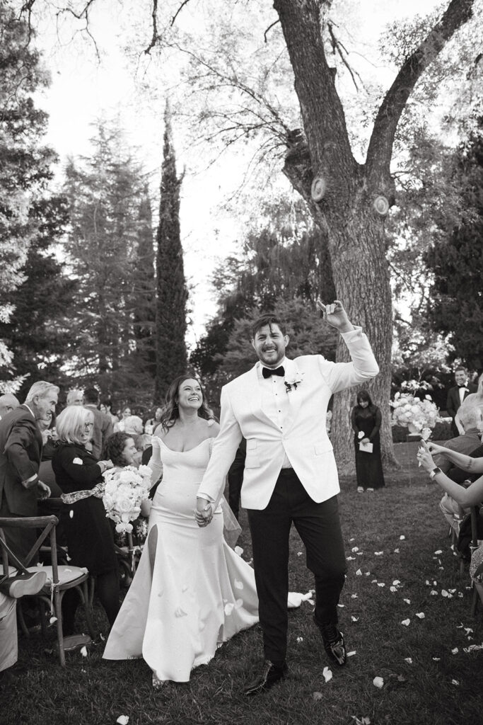 Couple walking back down the aisle laughing and cheering while guests throw white rose petals.