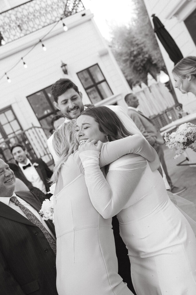 Bride tearfully hugging another woman as groom smiles in background.