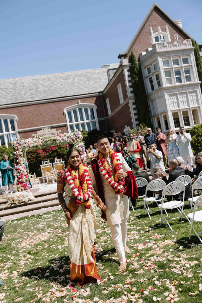 Multiracial couple walking down the aisle after a traditional Indian ceremony, adorned in red and yellow floral leis.